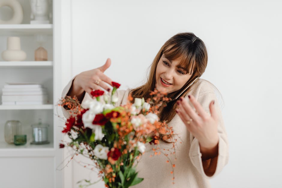 A woman with light brown hair and a beige sweater is holding a vibrant bouquet of fresh flowers while talking on a smartphone. The bouquet features a mix of red, white, and peach-colored blooms, inclu