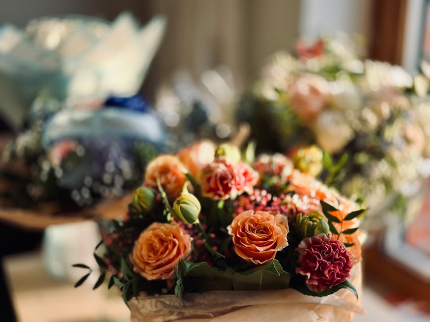 A floral arrangement featuring peach and coral roses, complemented by pink carnations and small white filler flowers, accented with green foliage and leaves. The bouquet is wrapped in light-colored pa