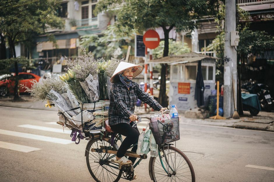 A woman wearing a traditional conical hat and a checkered shirt rides a bicycle along a city street. Attached to the bicycle's rear rack is a large, overflowing arrangement of fresh flowers, including