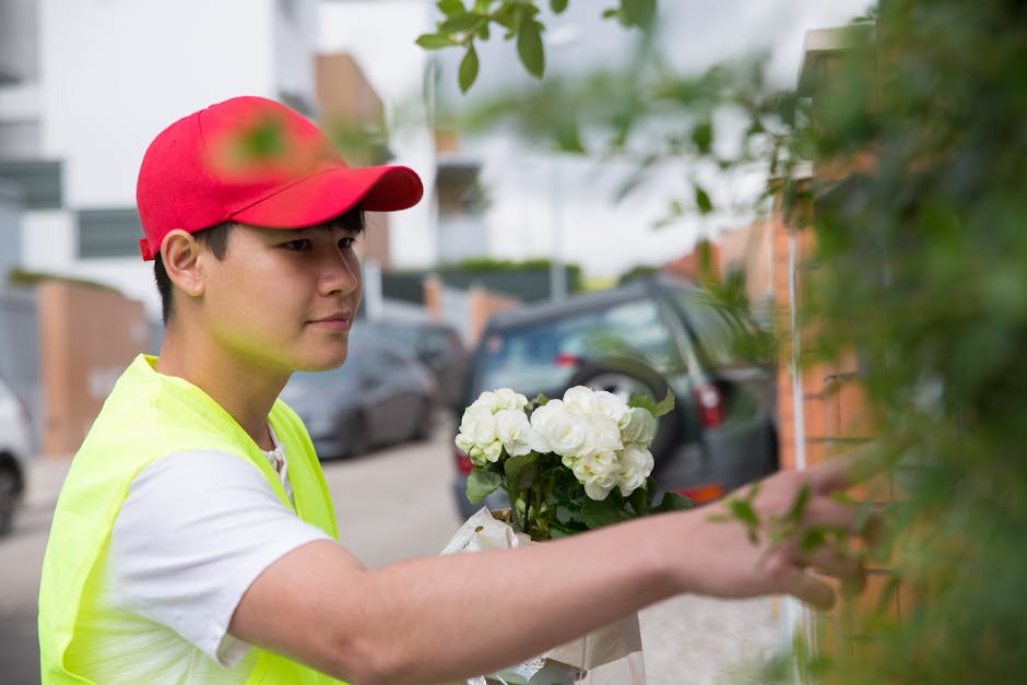 A delivery person wearing a red cap and a bright yellow high-visibility vest is seen handing over a bouquet of fresh, white hydrangeas wrapped in white paper, during a flower delivery. The flowers are