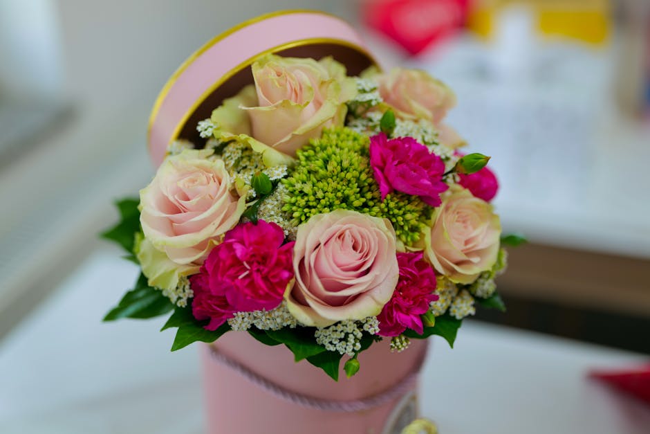 A round, pastel pink floral gift box contains a bouquet of fresh flowers, including soft pink roses with velvety petals, vibrant pink carnations, and clusters of small white and green flowers, arrange