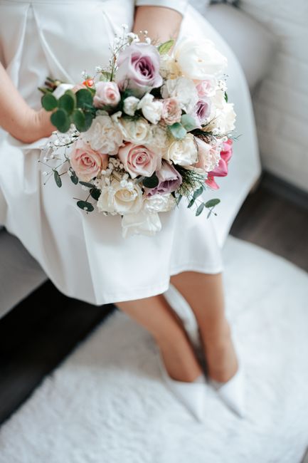 A bride appears seated on a white carpeted surface, holding an elegant bouquet of fresh flowers. The bouquet features soft pink, lavender, and white roses, accented with delicate baby's breath and lus