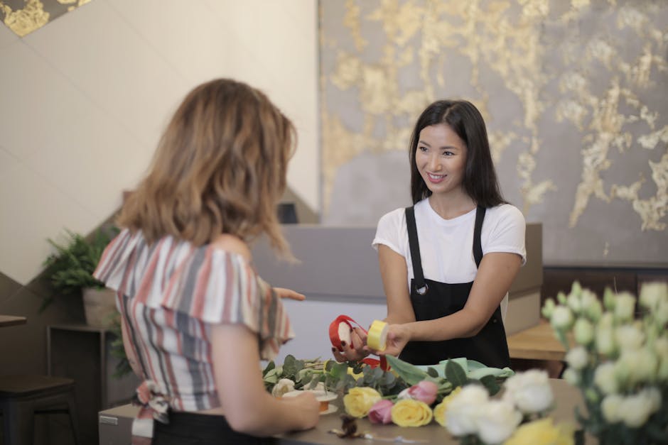 A floral shop employee with long dark hair, wearing a white t-shirt and black apron, hands a small, wrapped bouquet to a female customer with shoulder-length wavy blonde hair, dressed in a colorful st