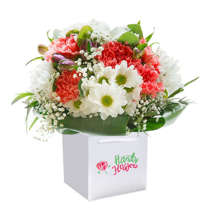 Presenting a flourish of joyous colour and soft texture, this premium floral bouquet stands upright in a square white carrier bag, poised for delivery to any Totteridge doorstep. The heart of the arrangement features generously frilled carnations in dreamy shades of coral, peach, and passionate red-their petals layered for depth and a touch of theatrical grace. Darting between them, white chrysanthemum daisies crowd together, each sporting a radiant lime-green center, echoing the excitement of spring's arrival along Totteridge's famous High Road. Lacy clusters of baby's breath impart an airy composition, the tiny white blossoms floating like dew caught in the grass. Carefully tucked on the left, a few unopened buds tinged with purple and pink enrich the bouquet's palette, adding gentle intrigue and dimension. Framing the blooms, a collar of deep, glossy leaves provides both structure and a lush, natural backdrop, reflecting the village's gardens in full summer leaf. The white carrier bag boasts firm handles and a smart, clear label, signalling local craftsmanship and thoughtful presentation. Lighting is soft and clinical, focusing on every vibrant detail of petal, leaf, and stem. Designed for accessibility and impact, this arrangement marks birthdays, anniversaries, and all heartfelt occasions across Totteridge, a testament to the care and flair of local N20 florists.