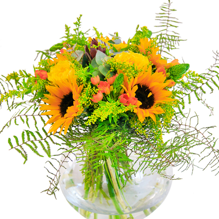 This sun-filled bouquet captures the spirit of Totteridge in late summer, blending radiant blooms with a touch of countryside charm. At its core are bright, cheerful sunflowers whose petal rays encircle rich, dark centers, summarising the optimism found in Totteridge's green open spaces. Clusters of luxurious yellow garden roses, their petals unfurling in generous spirals, nestle close by, lending a classic softness and a feast for the senses to the display. Glossy red Hypericum berries gleam among a cascade of fresh greenery: feathery ferns, lacy goldenrod, and slender flower spikes mimic the variety found along Dollis Valley Greenwalk. Subtle eucalyptus leaves introduce hints of blue-green, infusing a rustic element that balances the bold sunflowers. Arranged in a round, clear glass vase, every stem is visible, immersed in fresh water that accentuates the bouquet's vigour and staying power. The interplay of textures and colours-soft, suede-like petals; crisp, dewy foliage; vibrant berries-creates a lively composition. This arrangement evokes a carefree celebration, perfect for Totteridge homes, milestone occasions, or as a luminous centrepiece for family gatherings. It is meticulously crafted with local expertise, bringing the region's natural bounty to life and inviting joy into any room it graces.